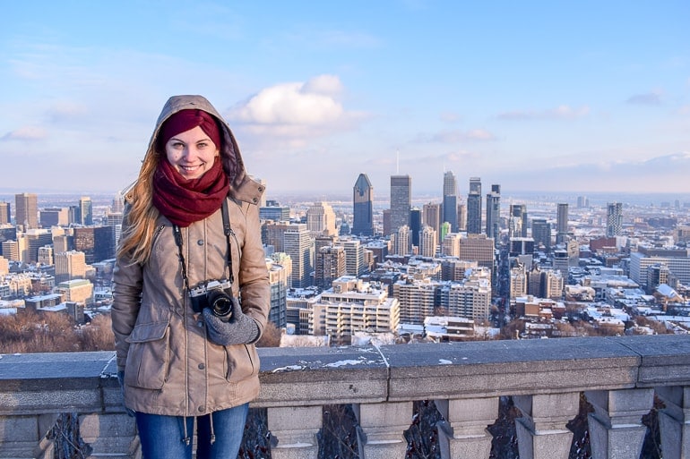 girl with tan jacket and red scarf holding camera with city behind.