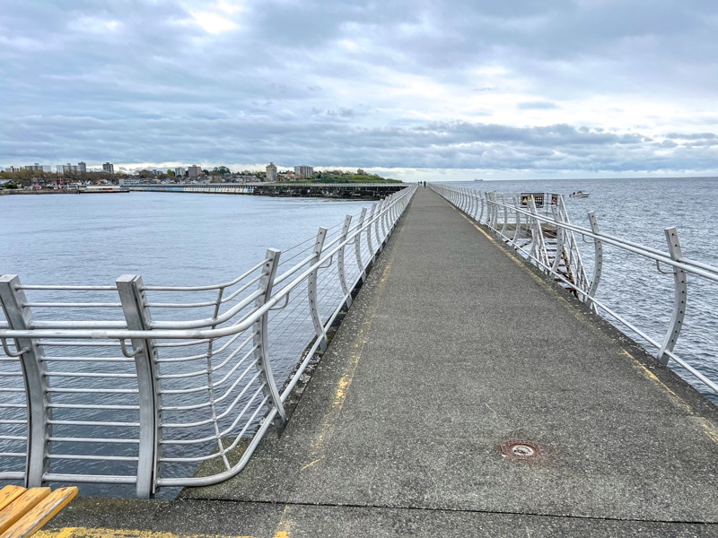long concrete walkway with metal railing and ocean on both sides and sky above.