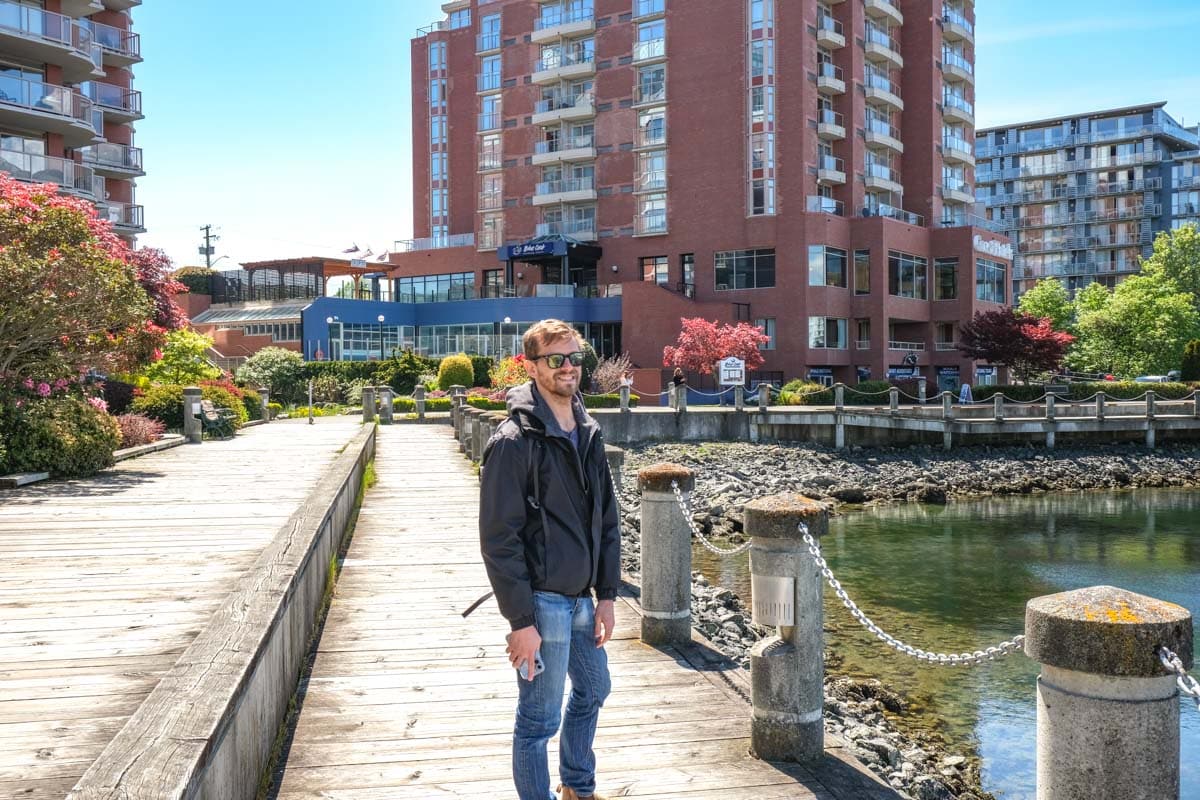 man with sunglasses walking along wooden boardwalk with harbour beside.