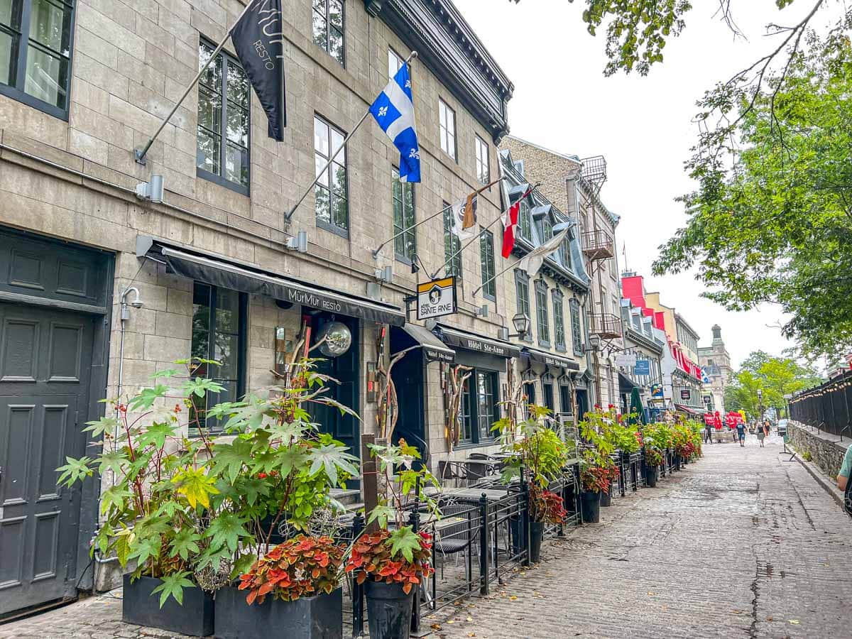 Historische Steingebäude mit Fahne über dem Bürgersteig und Pflanzen davor in der Altstadt Quebecs.
